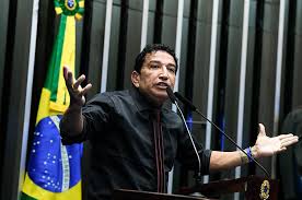 : Senador Magno Malta gesticulando durante sessão da CPI no Senado Federal, com microfone e bandeira ao fundo.