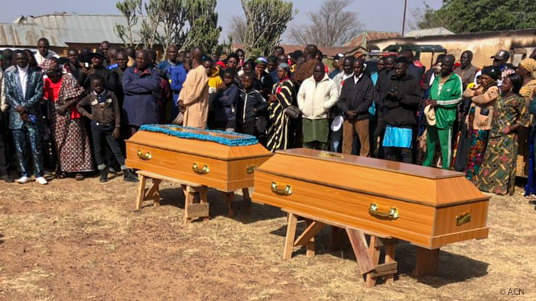 Multidão reunida em funeral ao ar livre com um padre discursando à frente de caixões.