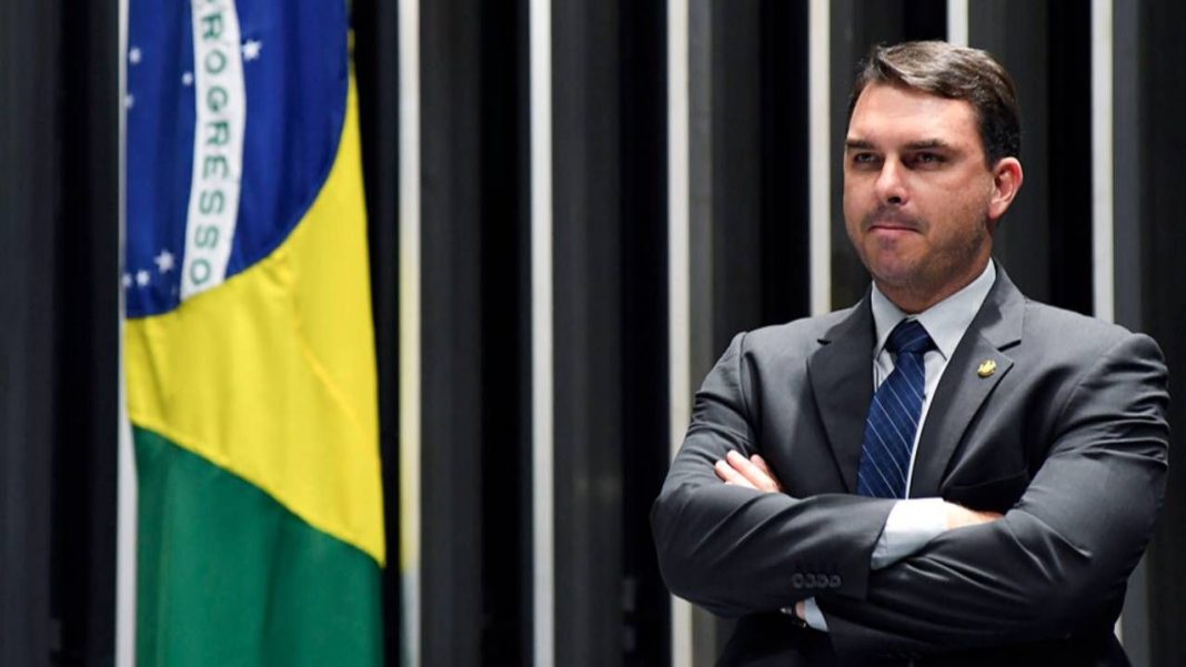 Foto do senador Flávio Bolsonaro discursando no Senado Federal com a bandeira do Brasil e o símbolo do TSE ao fundo em marca d'água.