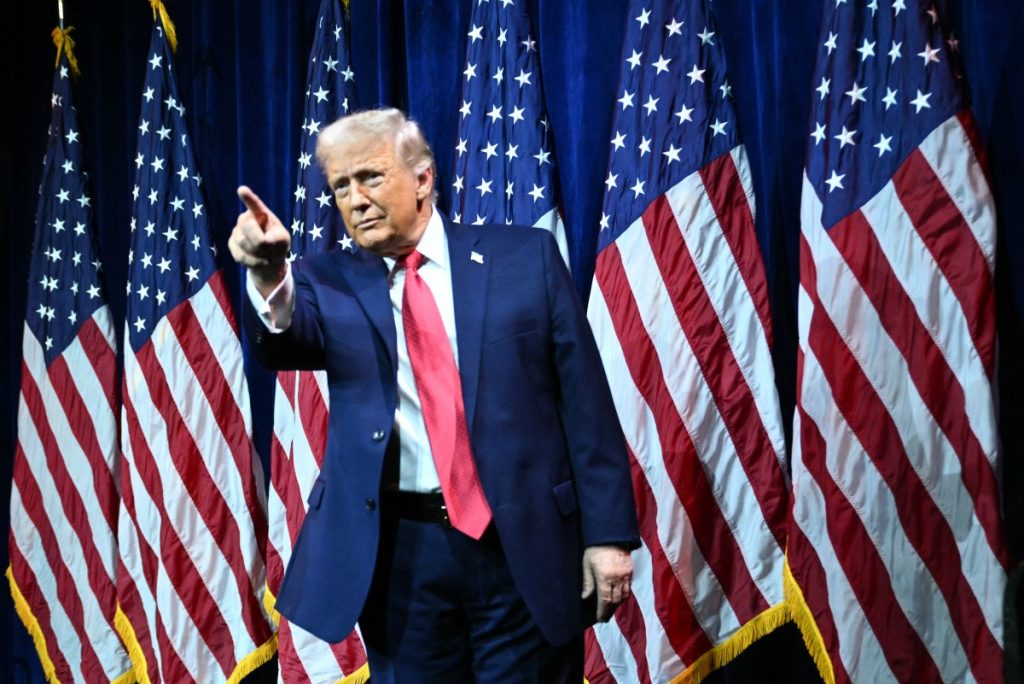 Donald Trump discursando em evento ao lado da bandeira dos Estados Unidos e da Venezuela estilizadas em fundo de crise diplomática.