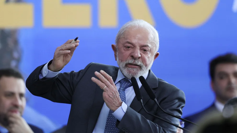 Presidente Lula discursando em palanque oficial em Itajaí vestindo casaco escuro e chapéu preto, com oficiais da Marinha ao fundo.